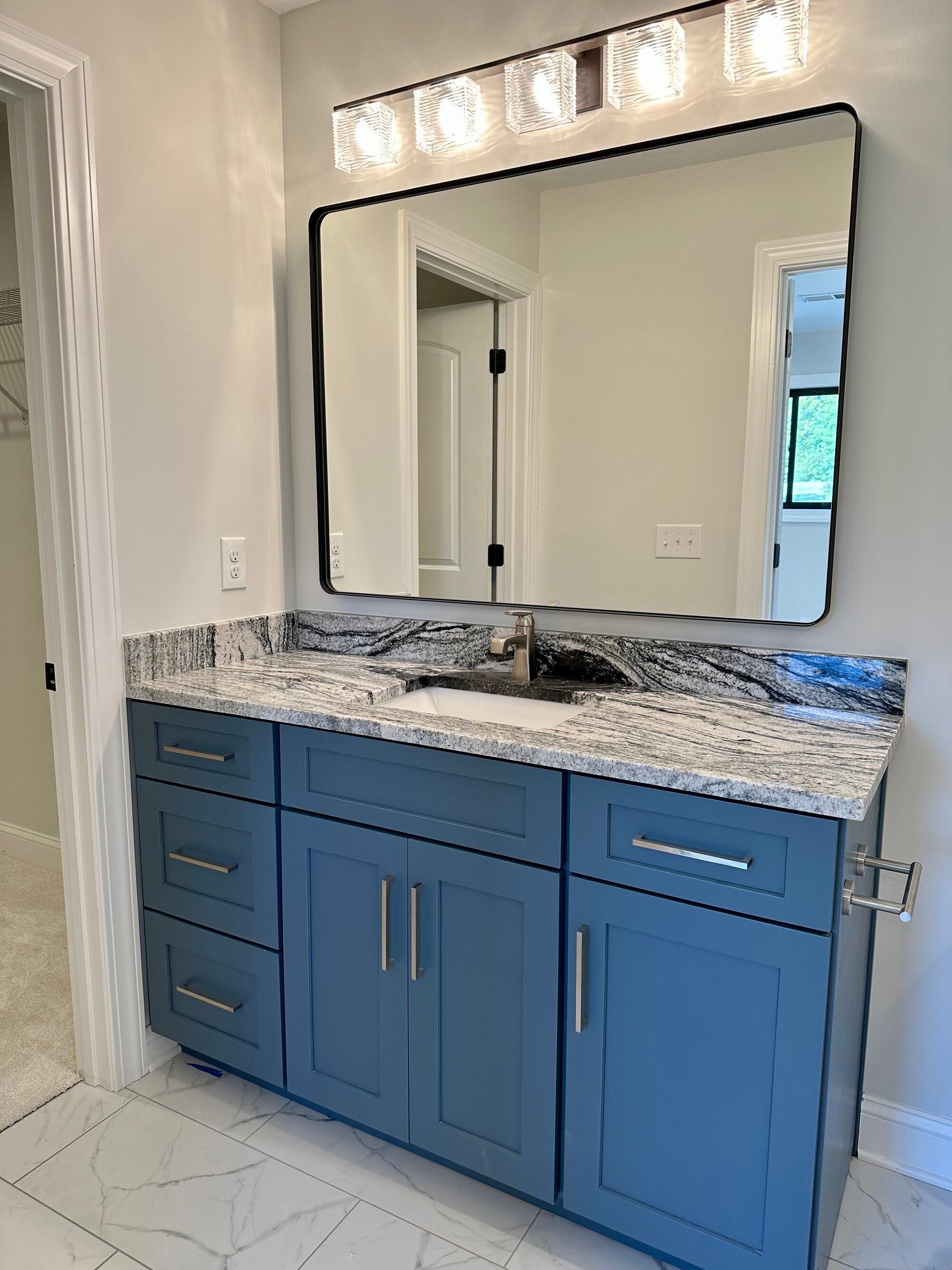 Bathroom with a large black-framed mirror, four modern lights, blue vanity with marbled countertop, silver handles, and white tile floor with gray veining